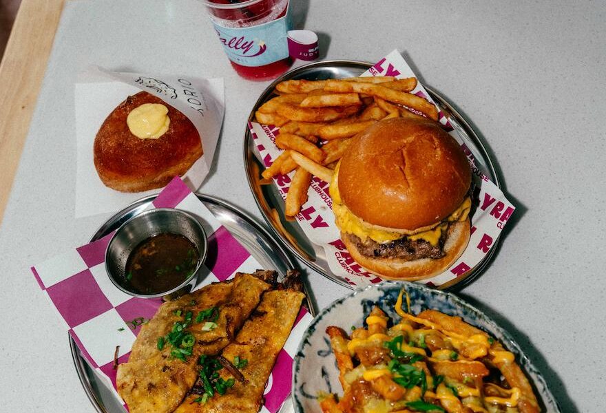 Overhead view of a Rally House table featuring a cheeseburger with fries, loaded fries, grilled street tacos with dipping sauce, a glazed doughnut, and two colorful beverages.