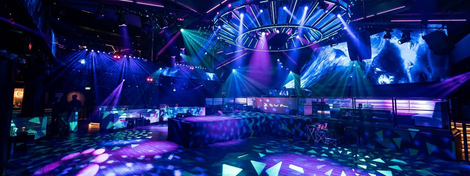 Wide view of Zouk's main room showcasing the central DJ booth, immersive LED displays, colorful stage lighting, spacious dance floor, and surrounding VIP lounge seating.