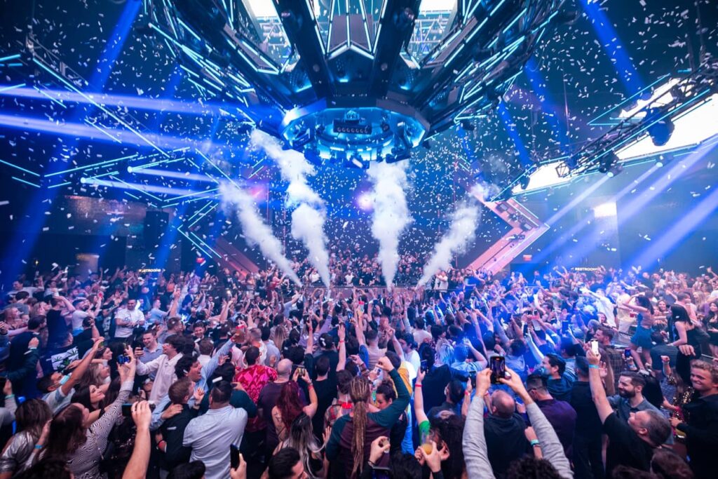 Crowd celebrating on a packed nightclub dance floor with confetti, stage lighting, and CO2 smoke effects.